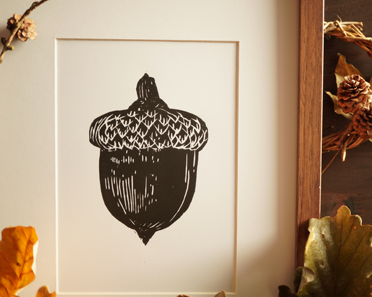 Linocut print of an acorn surrounded by autumn leaves.