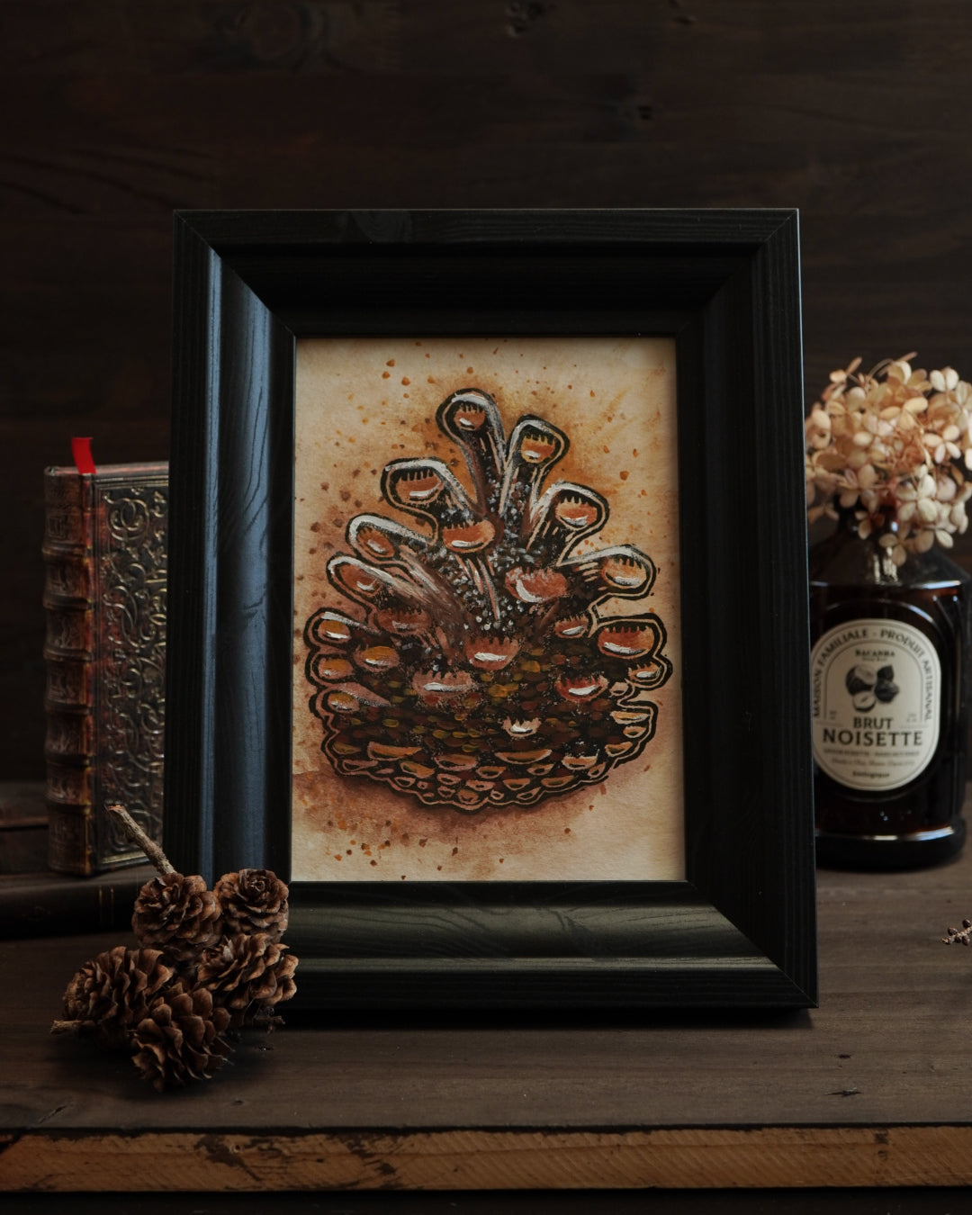Framed handmade linocut print of a pine cone by Hazel Lithwood, displayed with pinecones and forest-inspired objects.