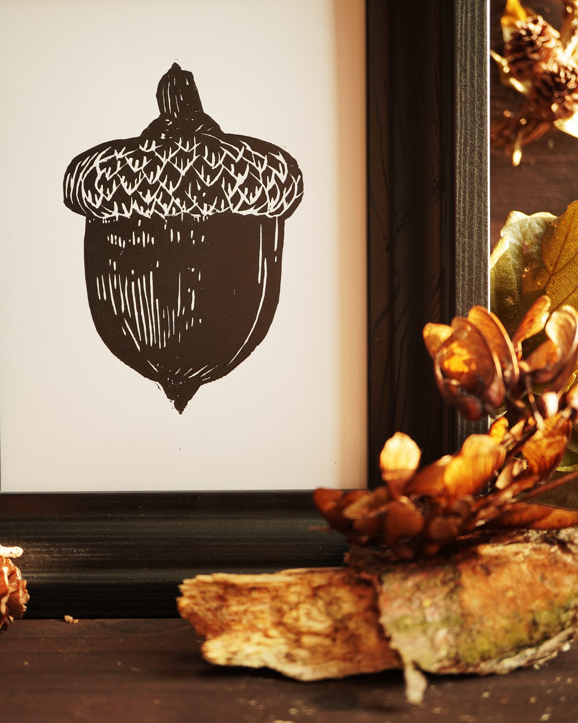 Linocut print of an acorn surrounded by autumn leaves.